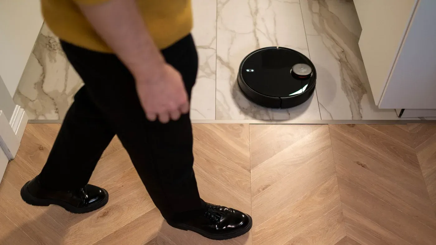 Svart robotdammsugare vid övergången mellan klinkergolv och fiskbensparkett medan en person går förbi